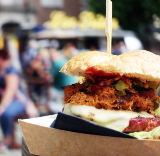 Ein appetitlicher Burger mit Schinken, Käse, Gemüse und einer würzigen Sauce auf einem Brötchen, serviert auf einem Papptablett vor einem unscharfen Hintergrund.